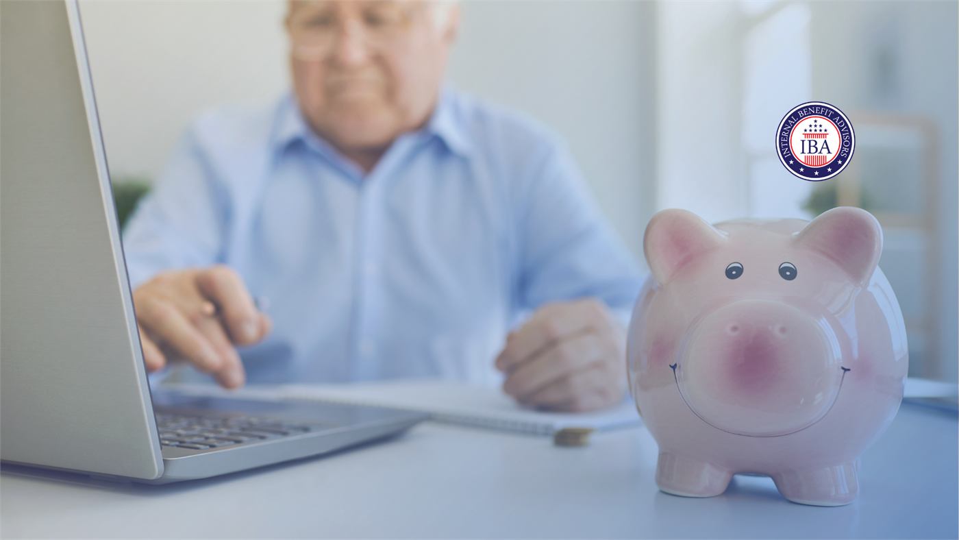 An older man working at a laptop at a white desk, with a ceramic piggy bank beside him