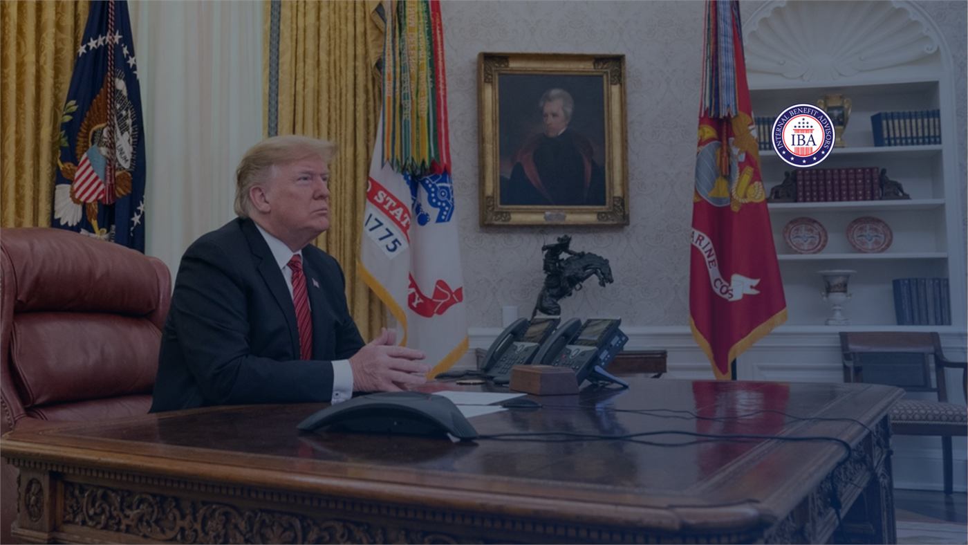 Donald Trump seated at the Resolute Desk in the Oval Office, with flags and a portrait behind him