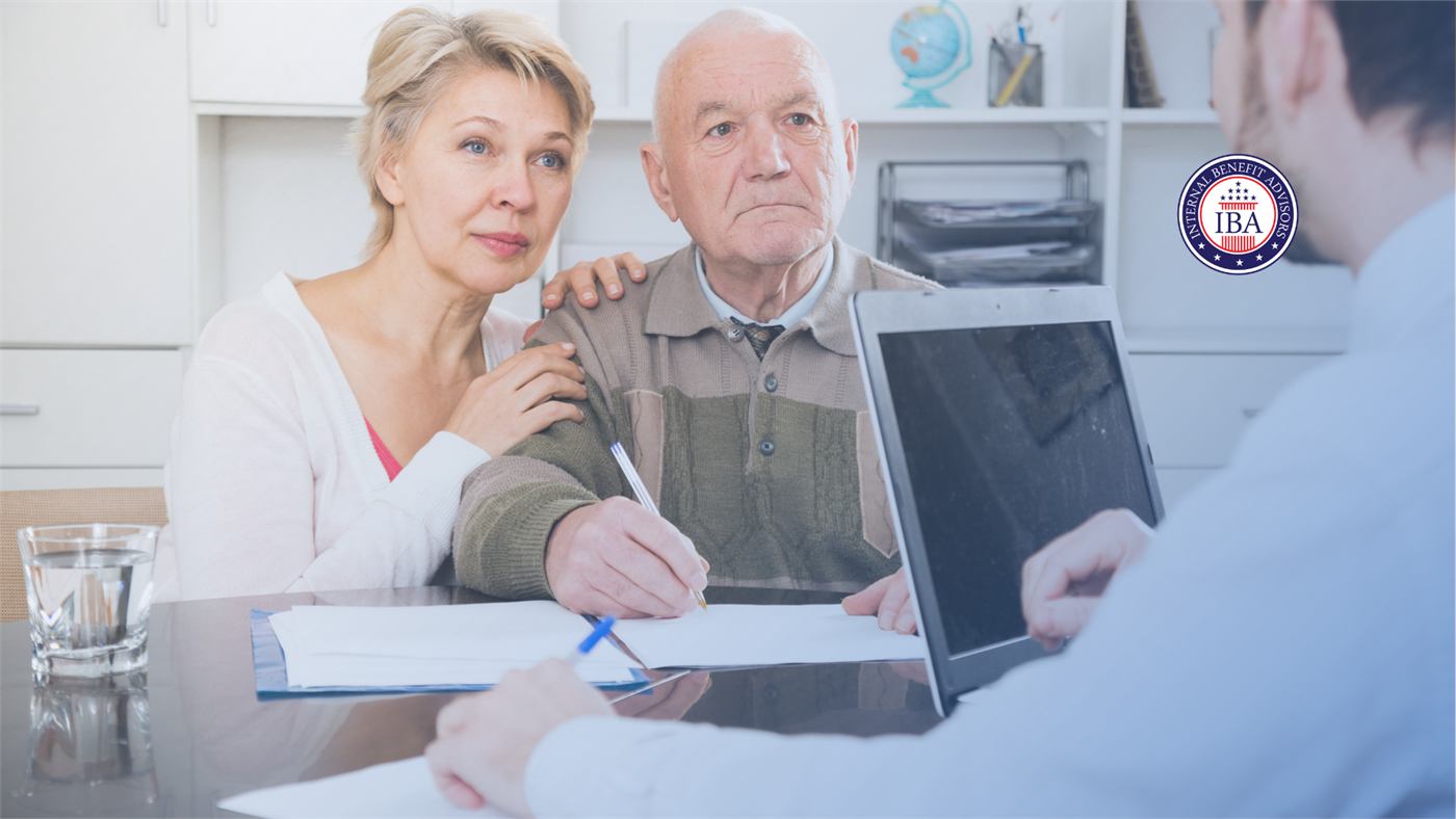 A woman and an older man signing paperwork at a desk while an adviser opposite points at a laptop screen