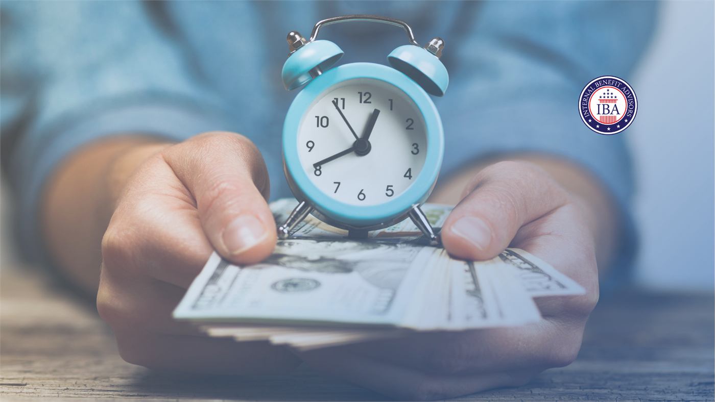 A pair of hands holding a small blue alarm clock above a fan of US banknotes