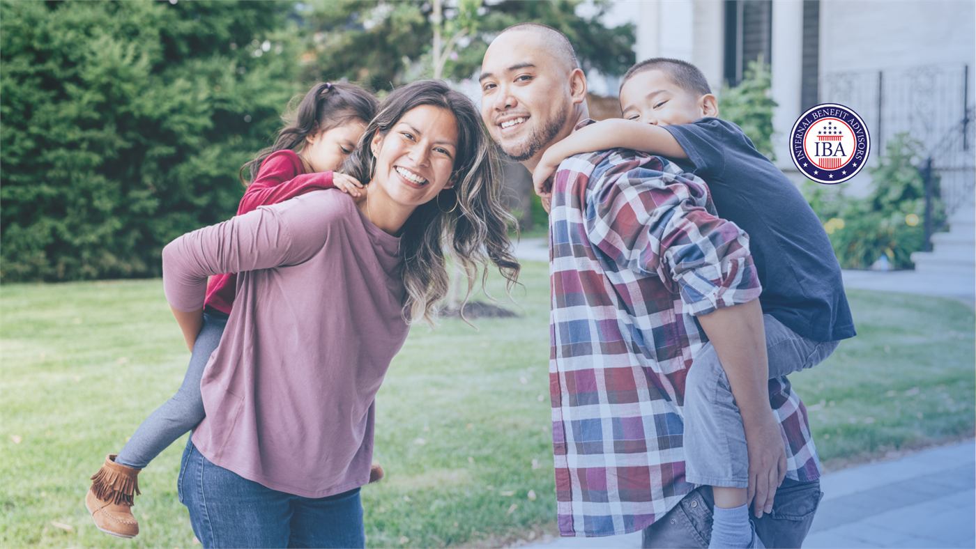 Two adults carrying children on their backs on a lawn outside a house, all of them laughing