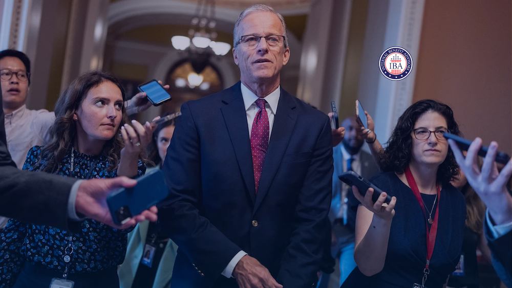 A man in a suit walking through a crowd of reporters holding phones and recorders in a marble corridor