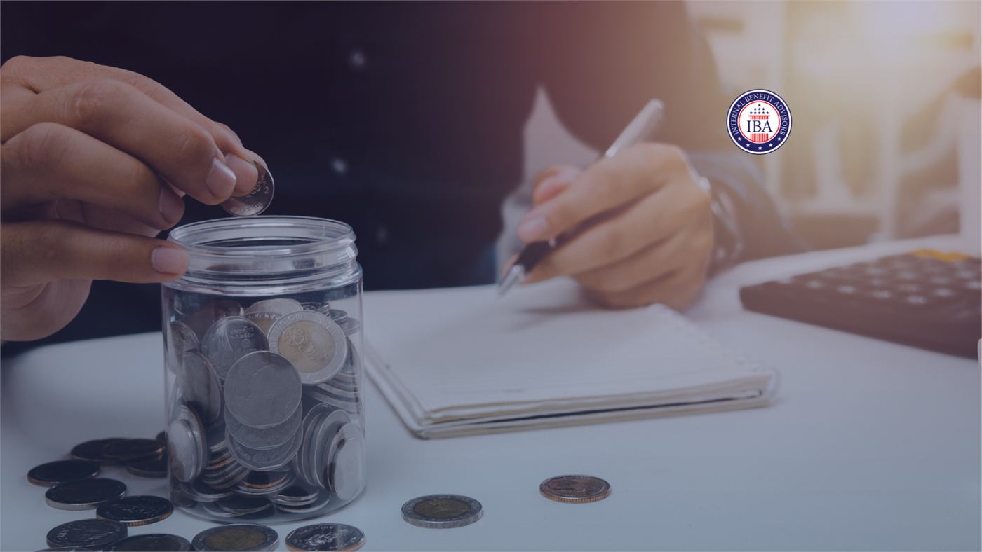 A hand dropping a coin into a glass jar of coins, beside a notebook, pen and calculator