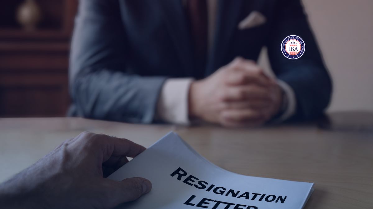 A man in a suit sitting at a desk with his hands clasped, a resignation letter in front of him