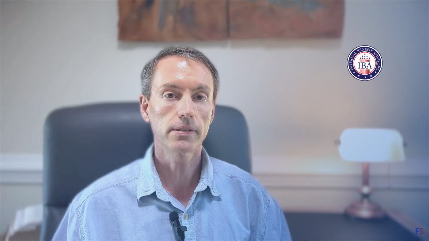 A man in a blue shirt speaking to camera in an office, a microphone in front of him