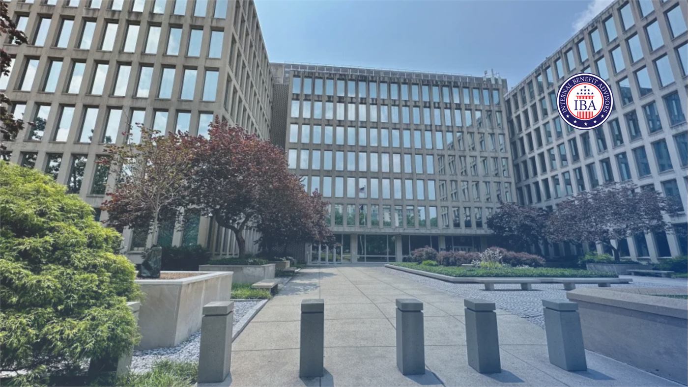 A modern government office building fronted by a wide paved plaza and low shrubs