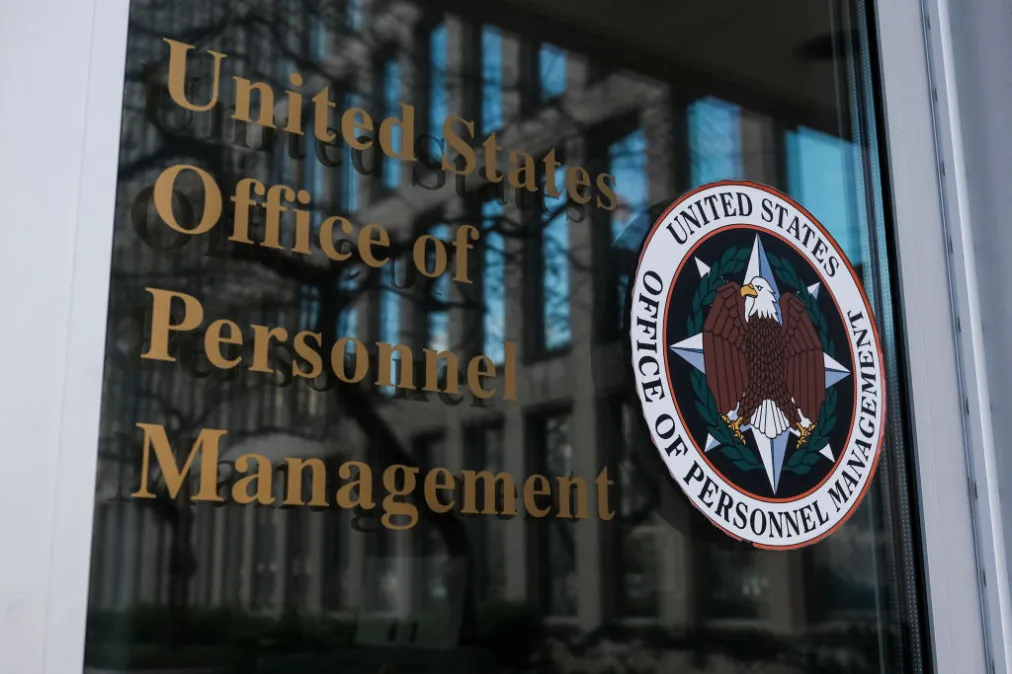 The United States Office of Personnel Management name and seal etched on a glass door, the building reflected behind