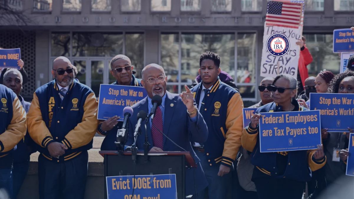 A man speaking at a press conference surrounded by union members holding signs reading Stop the War on America's Workforce and Federal Employees Are Tax Payers Too