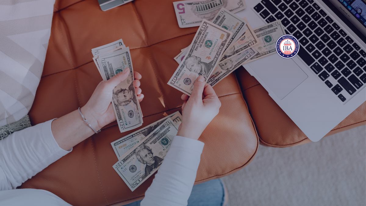 A person counting US banknotes on a sofa beside an open laptop