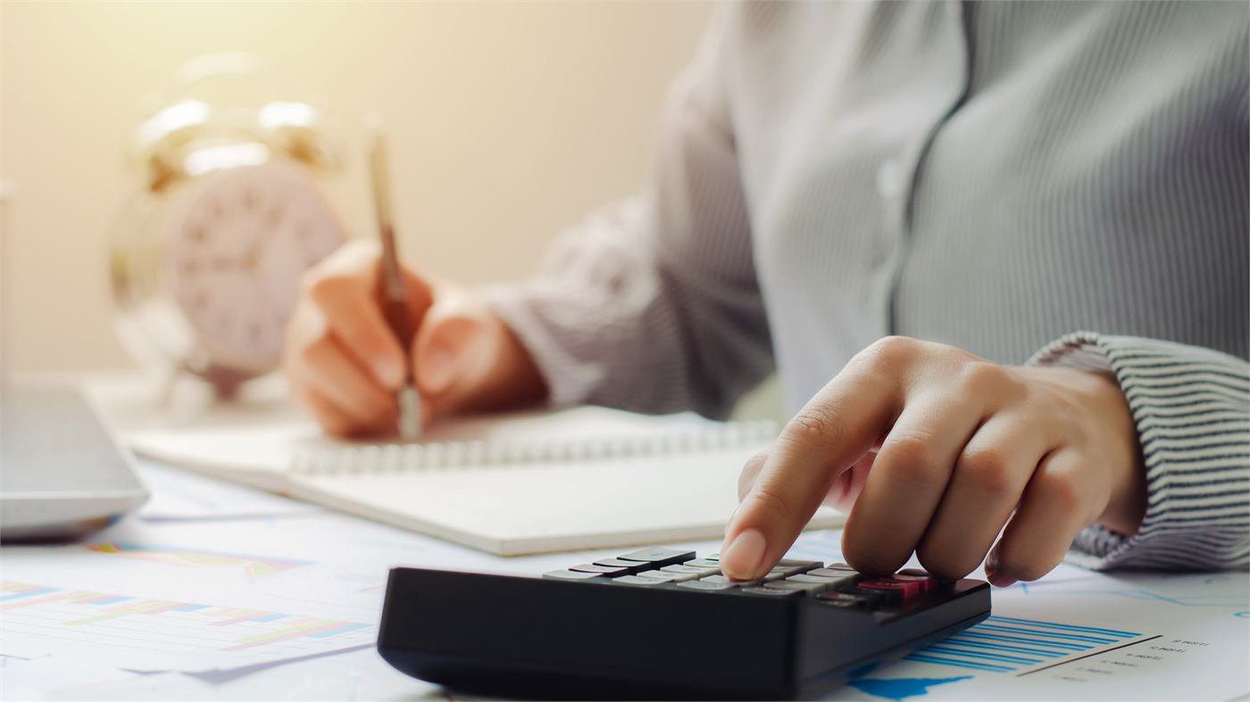 Hands using a calculator beside a notebook and printed charts, an alarm clock in the background