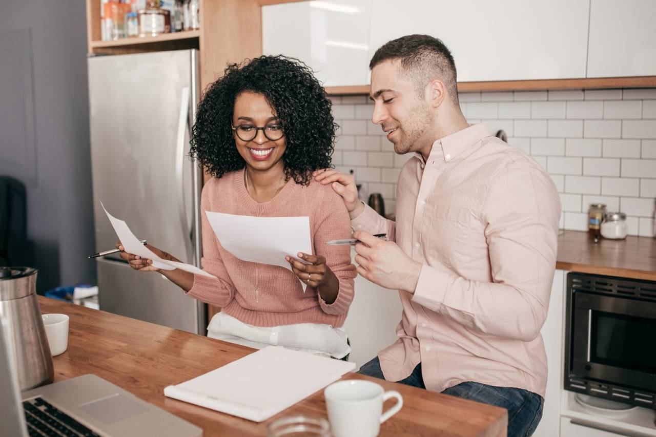 A couple in a kitchen reading printed documents together beside a laptop