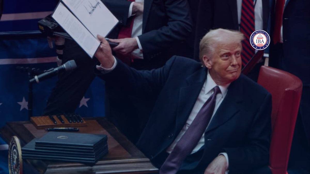 Donald Trump seated at a desk as an aide holds up a signed document