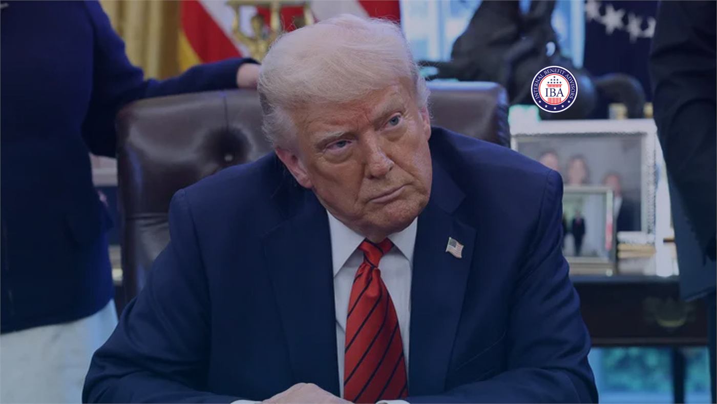 Donald Trump seated at the Resolute Desk with aides standing behind him
