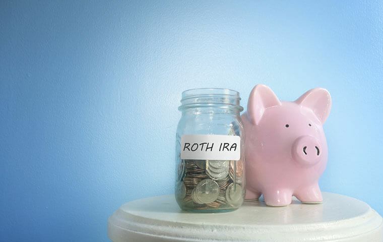A glass jar labelled Roth IRA filled with coins, beside a pink piggy bank